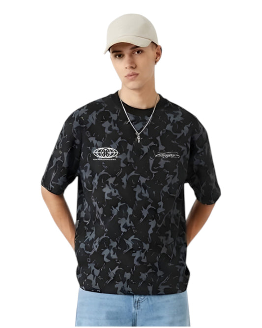 Anetos Blackout Camo Tee with a relaxed fit, featuring a black and grey camouflage pattern and short sleeves. Model wears light wash jeans and a beige cap.